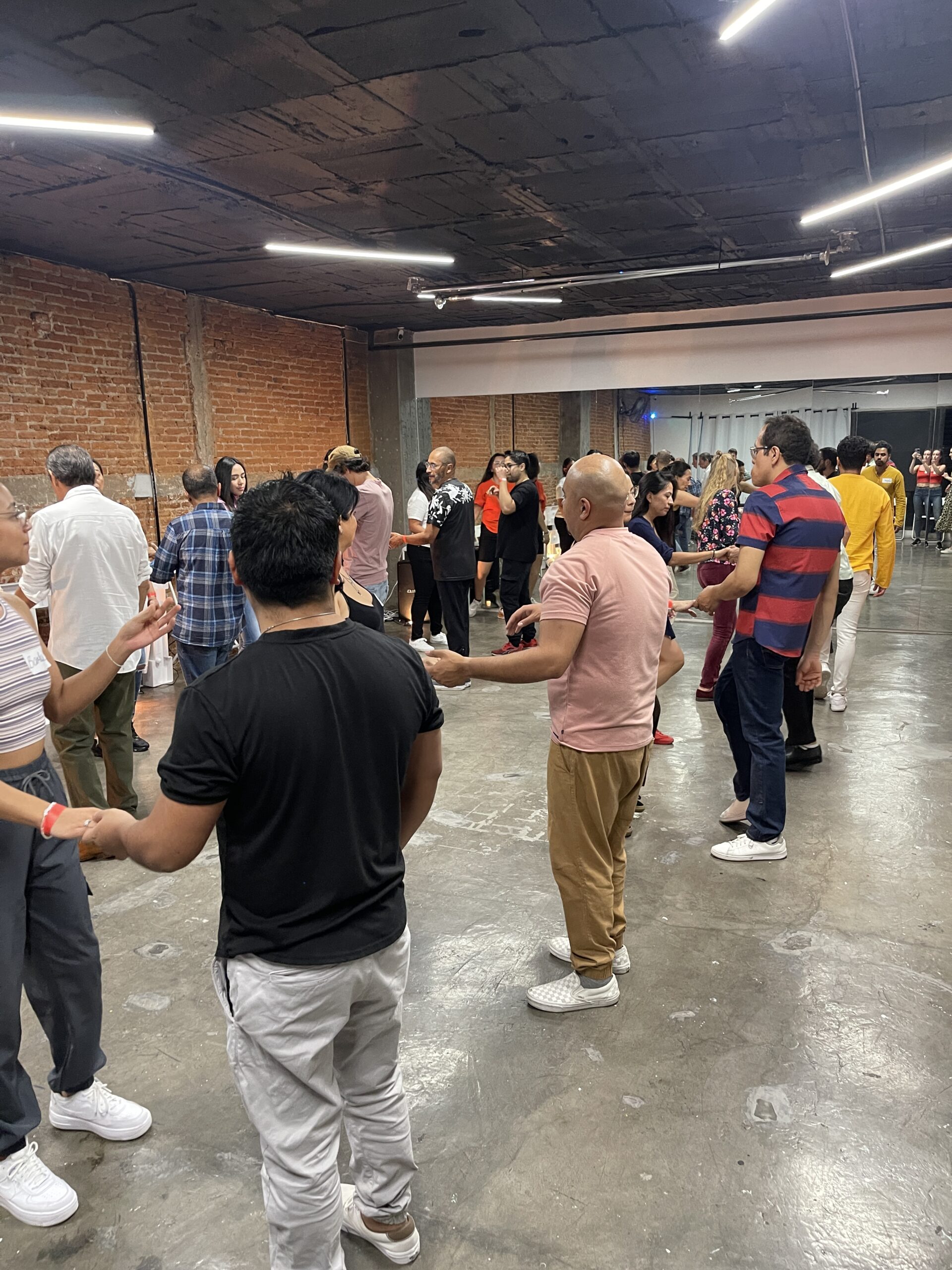 A group of beginner West Coast Swing students dancing in pairs inside a modern studio with exposed brick walls and concrete floors. The room is lively, with dancers facing each other and practicing basic steps under bright ceiling lights.